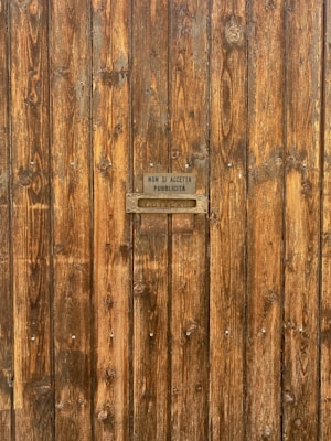 A wooden door with a textured surface, featuring vertical planks and a small metal sign. The sign reads 'Non si accetta pubblicit&agrave;' above a metal letter slot inscribed with 'Lettere'. The wood has a natural, rustic appearance with visible grain patterns and knots.