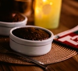 A warmly lit table setting featuring a white ramekin containing a chocolate dessert. A metal fork rests beside it on a woven placemat. In the background, there is a softly glowing candle inside a frosted glass holder. Partially visible, there is also a red-covered magazine.