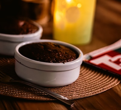A warmly lit table setting featuring a white ramekin containing a chocolate dessert. A metal fork rests beside it on a woven placemat. In the background, there is a softly glowing candle inside a frosted glass holder. Partially visible, there is also a red-covered magazine.