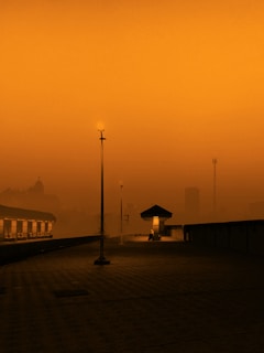 A solitary, dimly lit train station platform at dusk enveloped in an orange haze. A single light pole illuminates the deserted platform, while faint silhouettes of buildings and a radio tower appear in the background. The atmosphere evokes an otherworldly and tranquil ambiance.