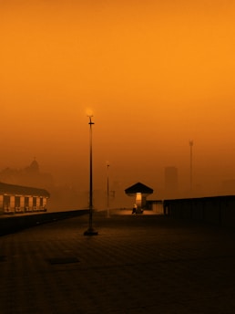 A solitary, dimly lit train station platform at dusk enveloped in an orange haze. A single light pole illuminates the deserted platform, while faint silhouettes of buildings and a radio tower appear in the background. The atmosphere evokes an otherworldly and tranquil ambiance.