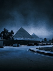 Two large pyramids are seen in the background of a night scene, illuminated in a bluish tone. In the foreground, a quiet street curves, and there are several parked cars alongside a wall. Trees and some vegetation are visible to the side.