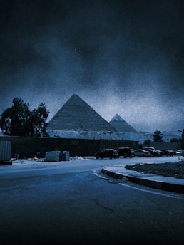 Two large pyramids are seen in the background of a night scene, illuminated in a bluish tone. In the foreground, a quiet street curves, and there are several parked cars alongside a wall. Trees and some vegetation are visible to the side.