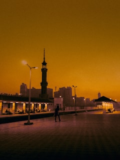 A train platform at dusk with a silhouette of a tall, ornate tower and a few people scattered across the platform. The sky is a striking orange, casting a warm glow over the scene. Streetlights are on, and some buildings in the background create an urban atmosphere.