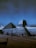 A majestic view of the Egyptian Sphinx and a pyramid under a dark sky with a crescent moon visible. There are a few people in the foreground, likely tourists, taking in the historical sights. The image captures the monumental structures in contrast to the serene night sky.