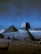 A majestic view of the Egyptian Sphinx and a pyramid under a dark sky with a crescent moon visible. There are a few people in the foreground, likely tourists, taking in the historical sights. The image captures the monumental structures in contrast to the serene night sky.