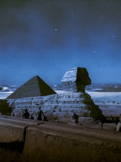 A majestic view of the Egyptian Sphinx and a pyramid under a dark sky with a crescent moon visible. There are a few people in the foreground, likely tourists, taking in the historical sights. The image captures the monumental structures in contrast to the serene night sky.