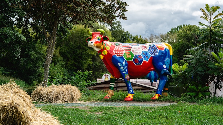 A vibrantly painted cow sculpture stands in a green park-like setting, featuring bold patterns and colors such as red, blue, and yellow. It is surrounded by lush greenery, hay bales, and trees, under a cloudy sky.