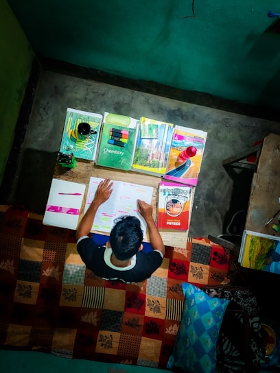 A person is sitting at a desk studying with several textbooks and notes spread out in front of them. The books cover subjects like chemistry, biology, and physics. The desk is cluttered with pens, highlighters, and a small decorative object. The scene suggests a focused and studious atmosphere.