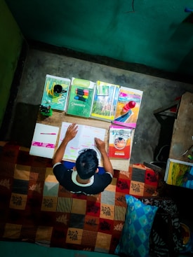 A person is sitting at a desk studying with several textbooks and notes spread out in front of them. The books cover subjects like chemistry, biology, and physics. The desk is cluttered with pens, highlighters, and a small decorative object. The scene suggests a focused and studious atmosphere.