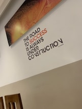 A motivational quote on a wall reads 'The road to success is always under construction.' The text is prominently displayed in black and orange, set against a light-colored wall. Above the quote, there is a rectangular artwork with a textured pattern in shades of orange and black. The bottom part of the room shows wooden chair backs.