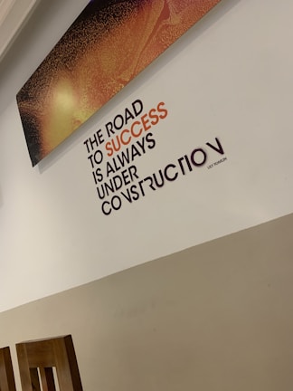 A motivational quote on a wall reads 'The road to success is always under construction.' The text is prominently displayed in black and orange, set against a light-colored wall. Above the quote, there is a rectangular artwork with a textured pattern in shades of orange and black. The bottom part of the room shows wooden chair backs.