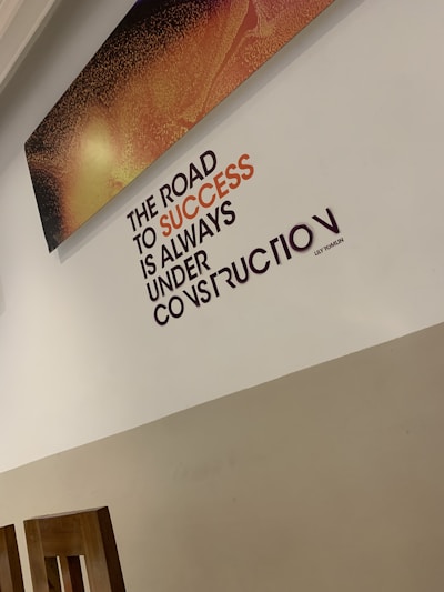 A motivational quote on a wall reads 'The road to success is always under construction.' The text is prominently displayed in black and orange, set against a light-colored wall. Above the quote, there is a rectangular artwork with a textured pattern in shades of orange and black. The bottom part of the room shows wooden chair backs.