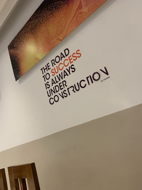 A motivational quote on a wall reads 'The road to success is always under construction.' The text is prominently displayed in black and orange, set against a light-colored wall. Above the quote, there is a rectangular artwork with a textured pattern in shades of orange and black. The bottom part of the room shows wooden chair backs.