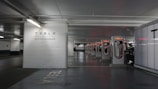 A row of Tesla Supercharger stations is situated in a dimly lit parking garage. The stations are aligned in a well-organized manner, with the Tesla logo and branding clearly visible. There is a car in the process of charging at one of the stations. The area appears clean and modern, with a clear focus on electric vehicle charging infrastructure.