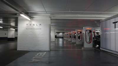 A row of Tesla Supercharger stations is situated in a dimly lit parking garage. The stations are aligned in a well-organized manner, with the Tesla logo and branding clearly visible. There is a car in the process of charging at one of the stations. The area appears clean and modern, with a clear focus on electric vehicle charging infrastructure.