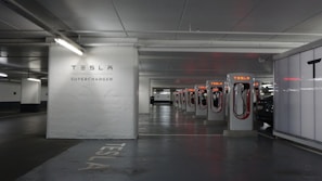 A row of Tesla Supercharger stations is situated in a dimly lit parking garage. The stations are aligned in a well-organized manner, with the Tesla logo and branding clearly visible. There is a car in the process of charging at one of the stations. The area appears clean and modern, with a clear focus on electric vehicle charging infrastructure.