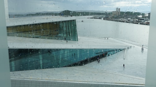 A modern architectural building with large glass windows is situated by a waterfront. People are walking on the large, flat roof of the building, which overlooks a calm body of water with boats. It is rainy, and you can see raindrops on the glass from which the photo is taken. Hills and an industrial area are visible in the background.