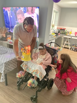 A young child seated in a wheelchair is surrounded by two adults, one of whom is holding a birthday cake with lit candles. The room has a television on the wall and a decorated table on the side. The child is wearing colorful and cozy clothing, including fluffy slippers.
