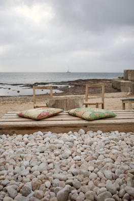 An outdoor setting with a stone-covered area leading to a wooden seating deck, equipped with colorful cushions. The background features a sandy shore with a calm sea and a small sailboat in the distance. The scene is under an overcast sky, giving it a serene and quiet atmosphere.