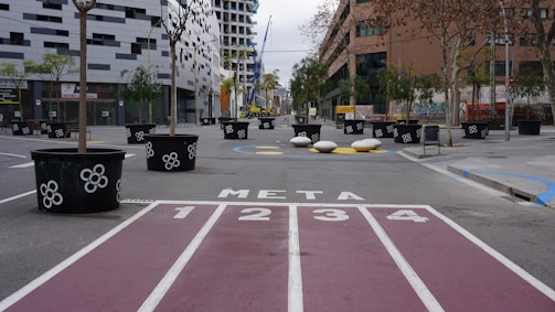 An urban setting featuring a pedestrian-friendly street with large black planters decorated with white floral patterns lining the road. The center displays a purple and white racing track-like path leading to a painted finish line marked 'META'. Modern buildings and bare-branched trees surround the open area.