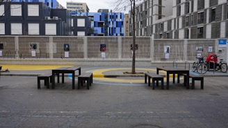 A modern, urban outdoor space with several black benches and tables, situated on a concrete surface with a tree planted in the center. Behind the benches, there is a wire fence displaying several posters. The background features a variety of contemporary buildings with a mix of gray and blue facades. There are a few bicycles parked on the right side.