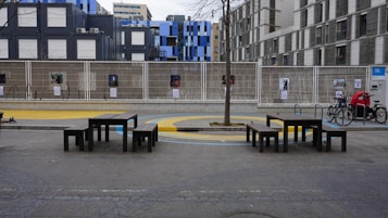 A modern, urban outdoor space with several black benches and tables, situated on a concrete surface with a tree planted in the center. Behind the benches, there is a wire fence displaying several posters. The background features a variety of contemporary buildings with a mix of gray and blue facades. There are a few bicycles parked on the right side.