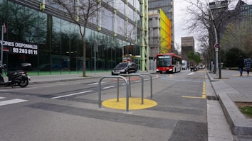 An urban street setting with a public bus and a few cars traveling down the road. There are glass-fronted buildings with bare trees lining the street. A scooter is parked nearby, and a couple of pedestrians are walking on the sidewalk. The road has various markings, and there are metallic barriers on a yellow circle near the center of the image.