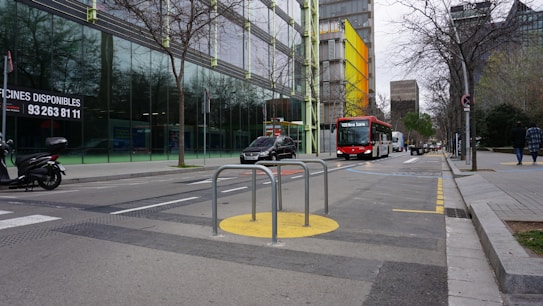 An urban street setting with a public bus and a few cars traveling down the road. There are glass-fronted buildings with bare trees lining the street. A scooter is parked nearby, and a couple of pedestrians are walking on the sidewalk. The road has various markings, and there are metallic barriers on a yellow circle near the center of the image.
