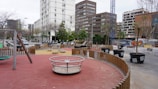 A modern urban playground is surrounded by tall buildings and a construction crane. The play area features equipment like a merry-go-round, swings, and seesaws, with pathways lined by small trees in large pots. There is a wooden fence segmenting the play zones, and beyond the playground, several scooters and motorcycles are parked along the street.