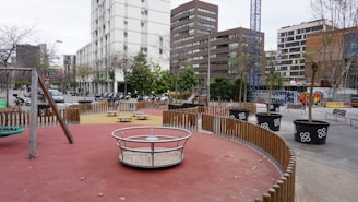 A modern urban playground is surrounded by tall buildings and a construction crane. The play area features equipment like a merry-go-round, swings, and seesaws, with pathways lined by small trees in large pots. There is a wooden fence segmenting the play zones, and beyond the playground, several scooters and motorcycles are parked along the street.