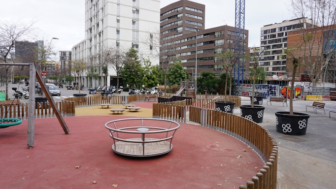 A modern urban playground is surrounded by tall buildings and a construction crane. The play area features equipment like a merry-go-round, swings, and seesaws, with pathways lined by small trees in large pots. There is a wooden fence segmenting the play zones, and beyond the playground, several scooters and motorcycles are parked along the street.