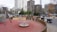 A modern urban playground is surrounded by tall buildings and a construction crane. The play area features equipment like a merry-go-round, swings, and seesaws, with pathways lined by small trees in large pots. There is a wooden fence segmenting the play zones, and beyond the playground, several scooters and motorcycles are parked along the street.