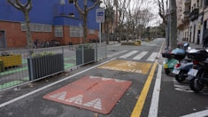 An urban street scene featuring a designated school zone with a yellow school crossing sign on the pavement. There are several motorcycles parked on one side and bicycles secured next to a building with blue and brick walls. Leafless trees line both sides of the street, while planters with greenery are placed at intervals along the sidewalk.