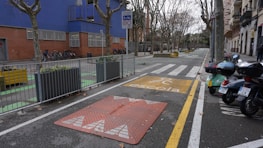 An urban street scene featuring a designated school zone with a yellow school crossing sign on the pavement. There are several motorcycles parked on one side and bicycles secured next to a building with blue and brick walls. Leafless trees line both sides of the street, while planters with greenery are placed at intervals along the sidewalk.