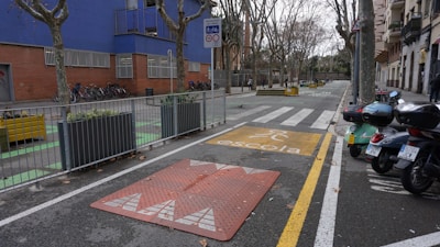 An urban street scene featuring a designated school zone with a yellow school crossing sign on the pavement. There are several motorcycles parked on one side and bicycles secured next to a building with blue and brick walls. Leafless trees line both sides of the street, while planters with greenery are placed at intervals along the sidewalk.