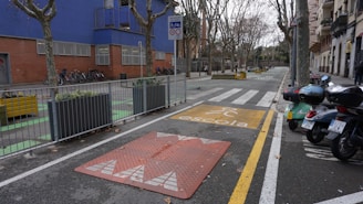 An urban street scene featuring a designated school zone with a yellow school crossing sign on the pavement. There are several motorcycles parked on one side and bicycles secured next to a building with blue and brick walls. Leafless trees line both sides of the street, while planters with greenery are placed at intervals along the sidewalk.