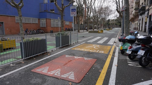 An urban street scene featuring a designated school zone with a yellow school crossing sign on the pavement. There are several motorcycles parked on one side and bicycles secured next to a building with blue and brick walls. Leafless trees line both sides of the street, while planters with greenery are placed at intervals along the sidewalk.