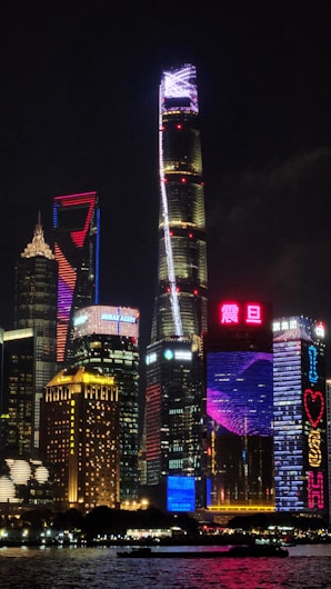 A stunning cityscape at night featuring a collection of skyscrapers illuminated with vibrant neon lights. The tallest building is highlighted by a vertical sequence of bright lights leading up to the top, complemented by surrounding structures with colorful display panels. Reflections shimmer on the water in the foreground, enhancing the lively atmosphere.