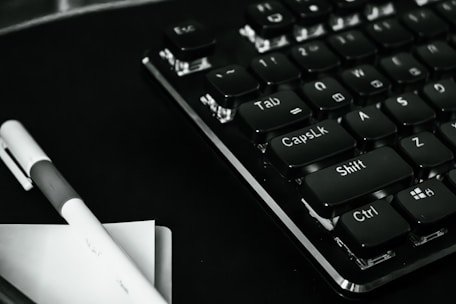 A close-up view of a black computer keyboard with prominently visible keys such as Tab, Caps Lock, Shift, Ctrl, and Esc. A pen and some papers are placed beside the keyboard on a dark surface, creating a monochrome look.