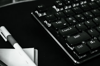 A close-up view of a black computer keyboard with prominently visible keys such as Tab, Caps Lock, Shift, Ctrl, and Esc. A pen and some papers are placed beside the keyboard on a dark surface, creating a monochrome look.