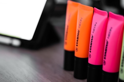 A close-up view of several neon-colored highlighters standing upright on a surface. The highlighters are in bright shades of pink and orange, with black bases, and are arranged in a line. An electronic device or screen is visible in the blurred background.