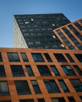 Tall buildings with modern architectural design are presented, showcasing a geometric pattern of windows. The structure is composed of different sections that slightly vary in color and texture, giving a dynamic and angled perspective against a clear blue sky.