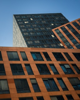 Tall buildings with modern architectural design are presented, showcasing a geometric pattern of windows. The structure is composed of different sections that slightly vary in color and texture, giving a dynamic and angled perspective against a clear blue sky.