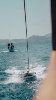 Two boats are on the open sea, with one towing the other. The water is a vivid shade of blue with visible ripples and white foam created by the movement of the boats. Hills are visible in the distant background under a clear sky.