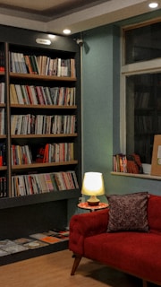 A cozy reading nook featuring a red upholstered couch with a patterned cushion next to a small side table holding a lit lamp. A large bookcase filled with books of various sizes and colors lines the wall. The lighting is warm, and the ambiance is inviting, suggesting a comfortable area for reading.