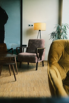 A cozy indoor scene featuring a plush armchair and a standing lamp with a warm yellow shade. The room has a parquet wooden floor, and there is a potted plant with long green leaves near the corner. A wooden side table with slender legs adds to the minimalist decor, while another armchair upholstered in a mustard fabric is partially visible in the foreground.