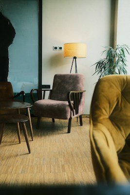 A cozy indoor scene featuring a plush armchair and a standing lamp with a warm yellow shade. The room has a parquet wooden floor, and there is a potted plant with long green leaves near the corner. A wooden side table with slender legs adds to the minimalist decor, while another armchair upholstered in a mustard fabric is partially visible in the foreground.