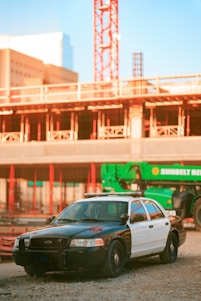 A police car is parked in front of a construction site. The structure in the background is partially built, with visible cranes and scaffolding. The police car is a black and white vehicle with visible roof lights, situated on gravel or dirt ground.