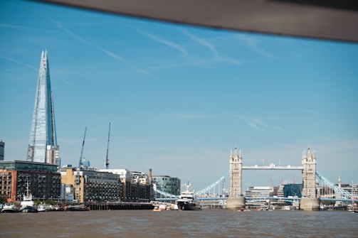 The image depicts a riverside cityscape featuring the iconic structure of a drawbridge with two towers. To the left, a tall, glass skyscraper stands prominently against a clear blue sky. Several construction cranes are visible in the background, along with various modern and historical buildings lining the waterfront.